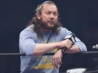 Kenny Omega blickt aus dem Ring und hält dabei ein Mikrofon in seiner rechten Hand