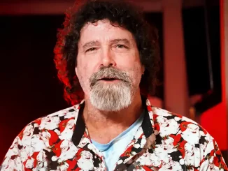 Mick Foley mit lockigem dunklem Haar und grau-weißem Vollbart blickt direkt in die Kamera, während er vor einem rot beleuchteten Hintergrund ein weißes Hemd mit auffälligem rot-schwarzem Muster trägt