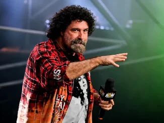 Mick Foley hält ein Mikrofon und erhebt seine Hand