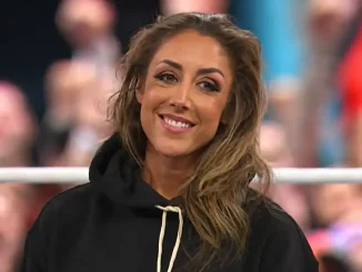 Die lächelnde Britt Baker mit den langen hellbraunen Haaren und dem schwarzen Hoodie steht vor einem verschwommenen Publikum und den Ringseilen einer Arena