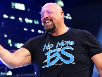 Paul Wight steht lachend in einer hell beleuchteten Wrestling-Arena und hält selbstbewusst eine geballte Faust in die Höhe
