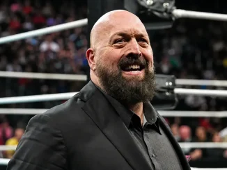 Paul Wight mit Vollbart steht vor einem Wrestling-Ring in einem dunklen Anzug, während im Hintergrund die Ringseile und eine verschwommene Zuschauermenge zu sehen sind