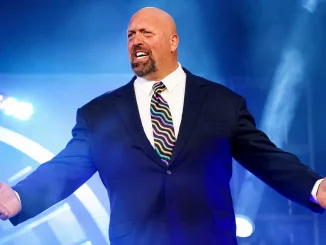 Paul Wight steht im dunklen Anzug und auffälliger Krawatte vor einer blau beleuchteten Wrestling-Bühne und breitet mit energischem Gesichtsausdruck beide Arme aus