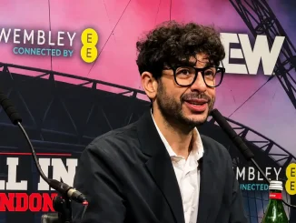 Tony Khan mit Brille und schwarzem Sakko sitzt während einer Pressekonferenz vor einer AEW-All-In-London-Kulisse mit Wembley-Schriftzügen und spricht in ein Mikrofon
