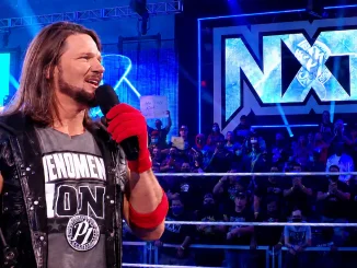 AJ Styles steht mit rotem Handschuh und Mikrofon im Ring, während hinter ihm das große, blau beleuchtete NXT-Logo und das Publikum zu sehen sind