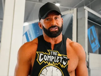 Matt Cardona mit schwarzer Kappe und Bart steht in einem hellen Fitnessstudio vor der Kamera und trägt ein schwarzes ärmelloses Shirt mit auffälligem „The Complete“-Motiv