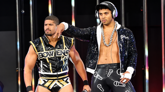Anthony Bowens und Max Caster stehen nebeneinander vor einer dunklen, beleuchteten Entrance-Kulisse; Bowens trägt sein schwarz-goldenes Ring-Outfit, während Caster mit schwarzer Paillettenjacke, Cap, Kopfhörern und auffälliger Silberkette posiert