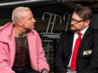 Auf dem Bild sitzen links Darby Allin in einer auffälligen pinkfarbenen Jacke und rechts Tony Schiavone im schwarzen Anzug mit roter Krawatte, während sich beide backstage einander zugewandt unterhalten