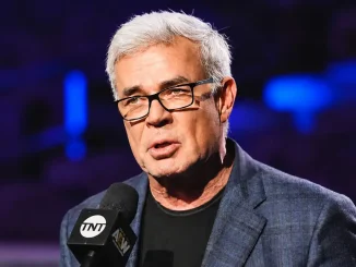 Eric Bischoff mit grauen Haaren und schwarzer Brille spricht in ein AEW-TNT-Mikrofon, während im Hintergrund unscharfe blau-violette Arena-Lichter zu sehen sind
