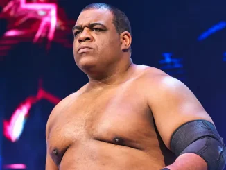 Auf dem Bild ist Keith Lee zu sehen. Er ist ein kräftig gebauter, oberkörperfreier Wrestler mit ernstem Gesichtsausdruck und schwarzer Ellenbogenbandage und steht vor einer farbig beleuchteten Arena-Kulisse