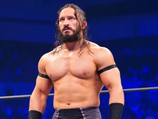 Die Grafik zeigt PAC mit langen dunklen Haaren und Bart, der mit ernstem Blick in einem blau beleuchteten Wrestling-Ring steht