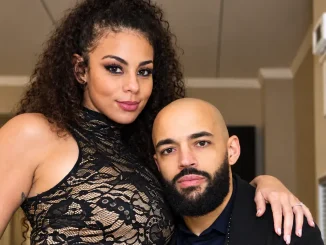 Zu sehen sind Samantha Irvin und Ricochet, die eng nebeneinander in einem hellen Innenraum posieren; Samantha trägt ein dunkles Outfit und legt ihren Arm auf Ricochets Schulter, während er in einem dunklen Hemd mit schwarzem Jackett in die Kamera blickt