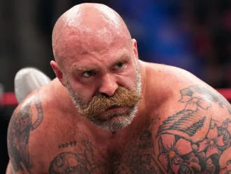 AEW-Wrestler The Butcher beugt sich mit angespanntem Blick nach vorn, wobei sein kahler Kopf, sein markanter Schnurrbart und die Tätowierungen auf seinem freien Oberkörper deutlich zu sehen sind