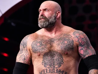 Andy „The Butcher“ Williams ist mit ernstem Blick und vollständig sichtbarem Kopf aus nächster Nähe zu sehen, wobei sein tätowierter Oberkörper vor einem dunklen Arena-Hintergrund im Mittelpunkt steht