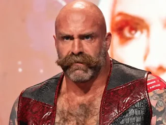AEW-Wrestler The Butcher blickt mit ernstem Gesichtsausdruck nach vorn und trägt eine offene rot-schwarze Weste, die seinen tätowierten Oberkörper teilweise freilässt