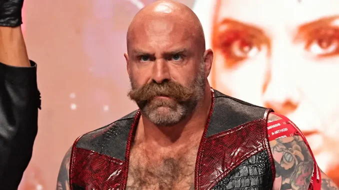 AEW-Wrestler The Butcher blickt mit ernstem Gesichtsausdruck nach vorn und trägt eine offene rot-schwarze Weste, die seinen tätowierten Oberkörper teilweise freilässt
