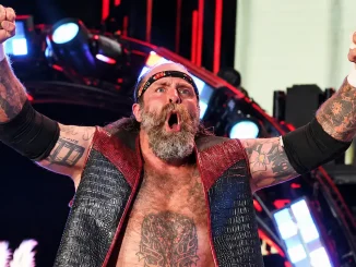 Andy Williams „The Butcher“ mit erhobenen Fäusten, geöffnetem Mund, markantem Vollbart, Stirnband, zahlreichen Tattoos, schwarzen Armschonern und einer schwarz-roten Weste vor einer hell beleuchteten Wrestling-Bühne mit roten und blauen Scheinwerfern
