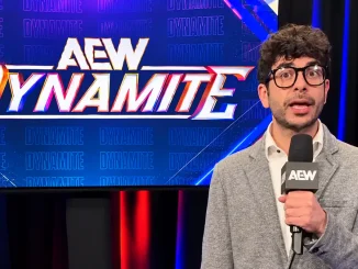 Tony Khan ist mit einem AEW-Mikrofon vor einem großen, vollständig sichtbaren AEW-Dynamite-Logo zu sehen