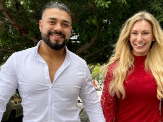Andrade im weißen Hemd und Charlotte Flair in einem roten Kleid gehen gemeinsam auf einem Gehweg unter grünen Bäumen entlang und blicken in die Kamera