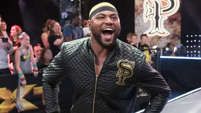 Angelo Dawkins ist gut gelaunt und mit weit geöffnetem Mund auf der WWE-Entrance-Stage zu sehen, während im Hintergrund Fans stehen und sein Logo auf der Videowand eingeblendet ist