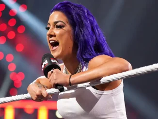 Bayley ist mit violetten Haaren im WWE-Ring zu sehen, während sie sich über die Ringseile lehnt und mit einem Mikrofon in der Hand lautstark zum Publikum spricht
