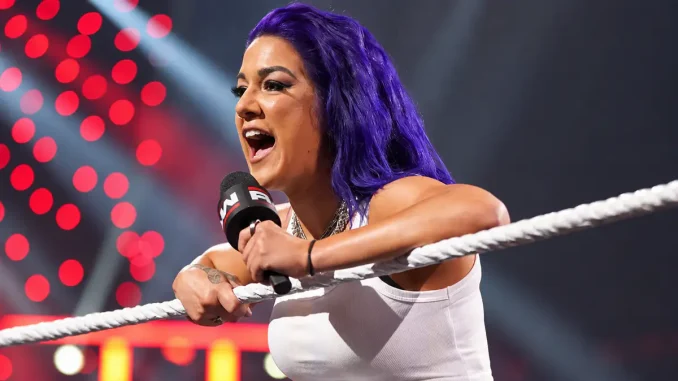 Bayley ist mit violetten Haaren im WWE-Ring zu sehen, während sie sich über die Ringseile lehnt und mit einem Mikrofon in der Hand lautstark zum Publikum spricht