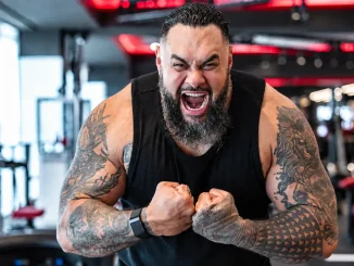 Bronson Reed ist in einem Fitnessstudio mit angespannten Armen, geballten Fäusten und lautem Schrei zu sehen, wobei seine kräftige Statur und die großflächigen Tattoos deutlich im Mittelpunkt stehen