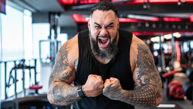Bronson Reed ist in einem Fitnessstudio mit angespannten Armen, geballten Fäusten und lautem Schrei zu sehen, wobei seine kräftige Statur und die großflächigen Tattoos deutlich im Mittelpunkt stehen