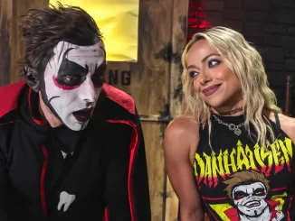 Danhausen und Liv Morgan stehen nebeneinander in einer düster inszenierten Backstage-Kulisse. Danhausen trägt seine markante schwarz-weiß-rote Gesichtsbemalung und einen schwarzen Umhang. Liv Morgan blickt lächelnd in einem schwarzen Danhausen-Shirt zu ihm