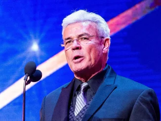 Eric Bischoff mit weißem Haar und Brille spricht in einem dunklen Anzug an einem Mikrofon vor einer blau beleuchteten Bühnenkulisse