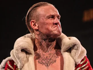 Ilja Dragunov ist mit ernstem, entschlossenem Gesichtsausdruck in einem auffälligen rot-goldenen Mantel mit Fellkragen zu sehen, während seine markanten Tattoos an Hals und Oberkörper deutlich hervorstechen