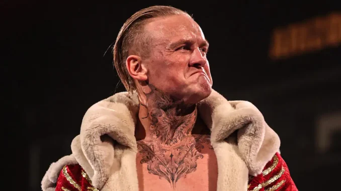 Ilja Dragunov ist mit ernstem, entschlossenem Gesichtsausdruck in einem auffälligen rot-goldenen Mantel mit Fellkragen zu sehen, während seine markanten Tattoos an Hals und Oberkörper deutlich hervorstechen