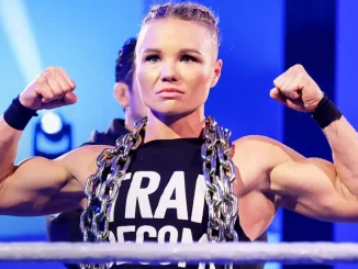 Zu sehen ist die muskulöse Wrestlerin Ivy Nile im Ring mit erhobenen, angespannten Armen und einer schweren Metallkette um den Hals vor einer blau beleuchteten Kulisse