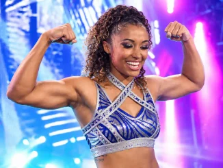 Kelani Jordan posiert lächelnd mit angespannten Armen vor einer hell beleuchteten Wrestling-Bühne und trägt dabei ein blau-silbernes, glitzerndes Ring-Outfit