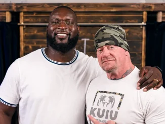 Omos und The Undertaker posieren gemeinsam vor einer Holzwand für die Kamera, während Omos lächelnd seinen Arm um die Schulter des Undertakers legt