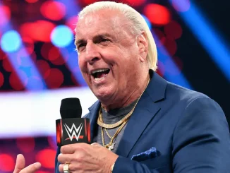 Ric Flair ist mit einem WWE-Mikrofon in der Hand zu sehen, während er lächelnd in einer Arena vor einem unscharfen rot-blau beleuchteten Hintergrund spricht