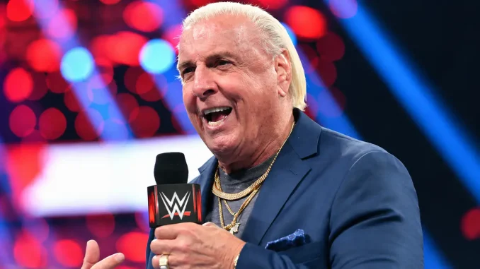 Ric Flair ist mit einem WWE-Mikrofon in der Hand zu sehen, während er lächelnd in einer Arena vor einem unscharfen rot-blau beleuchteten Hintergrund spricht