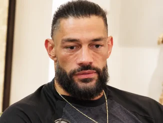 Roman Reigns ist in einer Nahaufnahme während eines Interviews zu sehen, mit ernstem, nachdenklichem Gesichtsausdruck, schwarzem Oberteil und goldener Halskette vor einem hellen Hintergrund