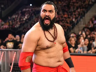 Sanga steht mit weit geöffnetem Mund und intensivem Gesichtsausdruck in roter Ringkleidung vor einer großen Zuschauerkulisse in einer Wrestling-Arena