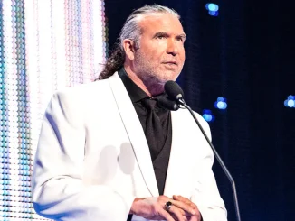 Scott Hall steht in einem weißen Anzug an einem Mikrofon auf einer hell beleuchteten Bühne und blickt mit ernster, konzentrierter Miene nach vorn