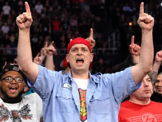 Wrestling-Fan Rick Achberger mit roter Kappe und blauem Hemd jubelt inmitten der Zuschauermenge mit beiden Armen und ausgestreckten Zeigefingern nach oben