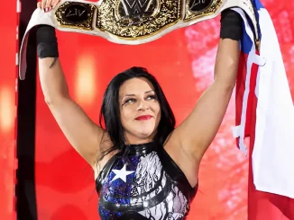 Stephanie Vaquer hält lächelnd einen WWE-Championship-Titel über ihren Kopf, während im Hintergrund eine rot beleuchtete Entrance-Bühne zu sehen ist