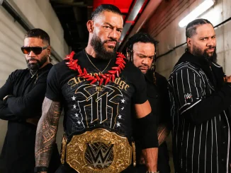 Das Bild zeigt vier schwarz gekleidete Männer der Bloodline in einem Backstage-Korridor, wobei der zentral stehende Mann mit roter Halskette und goldenem WWE-Championship-Gürtel selbstbewusst im Mittelpunkt steht