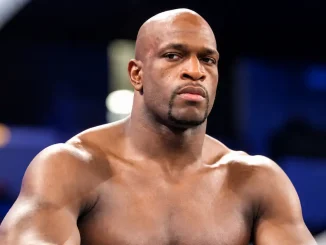 Titus O’Neil ist mit ernstem, fokussiertem Gesichtsausdruck und freiem Oberkörper vor dem unscharfen Hintergrund einer Wrestling-Arena zu sehen