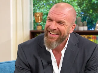 Zu sehen ist der glatzköpfige, vollbärtige Triple H in einem dunklen Anzug und weißem Hemd, der lächelnd auf einem blauen Sofa in einem hellen TV-Studio sitzt. Im Hintergrund befindet sich ein Regal mit Büchern, Dekorationen, Pflanzen und Blumen