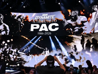 Das Bild zeigt die große AEW-Dynamite-Bühne mit einer eindrucksvollen Tribute-Grafik für einen verstorbenen Wrestler, während Zuschauer im Vordergrund auf die beleuchtete Videowand blicken.