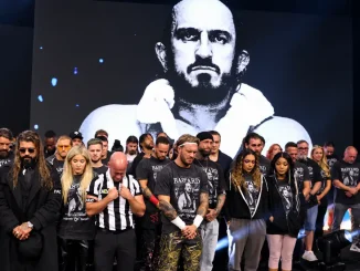 Zu sehen ist ein stiller Gedenkmoment bei AEW Dynamite für den verstorbenen Wrestler PAC: Zahlreiche Wrestlerinnen, Wrestler und Mitarbeiter stehen mit gesenkten Köpfen vor einem großen Schwarz-Weiß-Porträt von PAC, viele von ihnen tragen schwarze „BASTARD“-Tribute-Shirts.