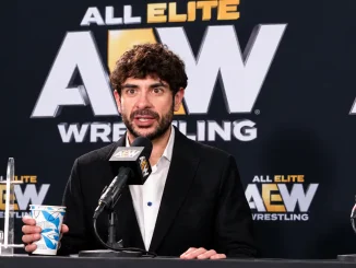 Tony Khan spricht bei einer AEW-Pressekonferenz in ein Mikrofon und hält einen Becher in der Hand. Im Hintergrund sind mehrere AEW-Logos zu sehen