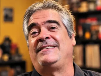Vince Russo mit grau meliertem Haar und Schnurrbart lächelt vor einem unscharfen Hintergrund in die Kamera.