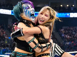 Asuka und Kairi Sane umarmen sich lachend im WWE-Ring. Die linke Wrestlerin Asuka trägt auffälliges blau-weißes Facepaint und türkis-violette Haare, während die blonde Wrestlerin Kairi sie freudestrahlend umarmt. Im Hintergrund sind die Zuschauer und die beleuchtete Arena zu sehen.